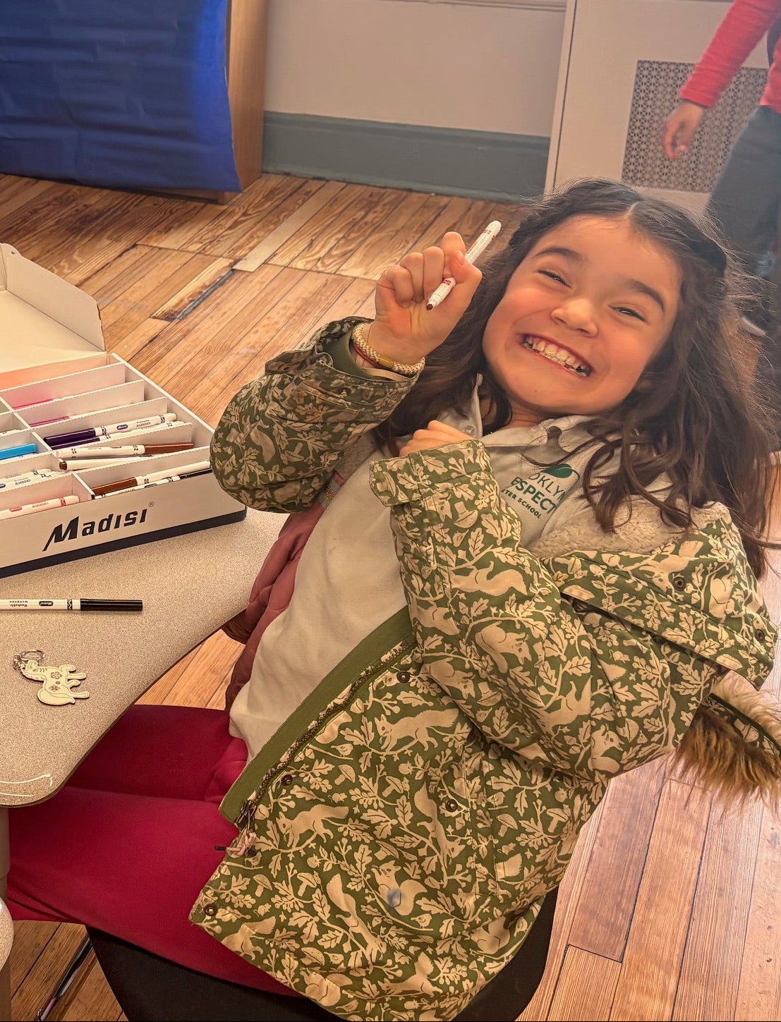 A student with a huge smile on her face sitting at a desk with a box of markers ready to color and customize her horse keychain.