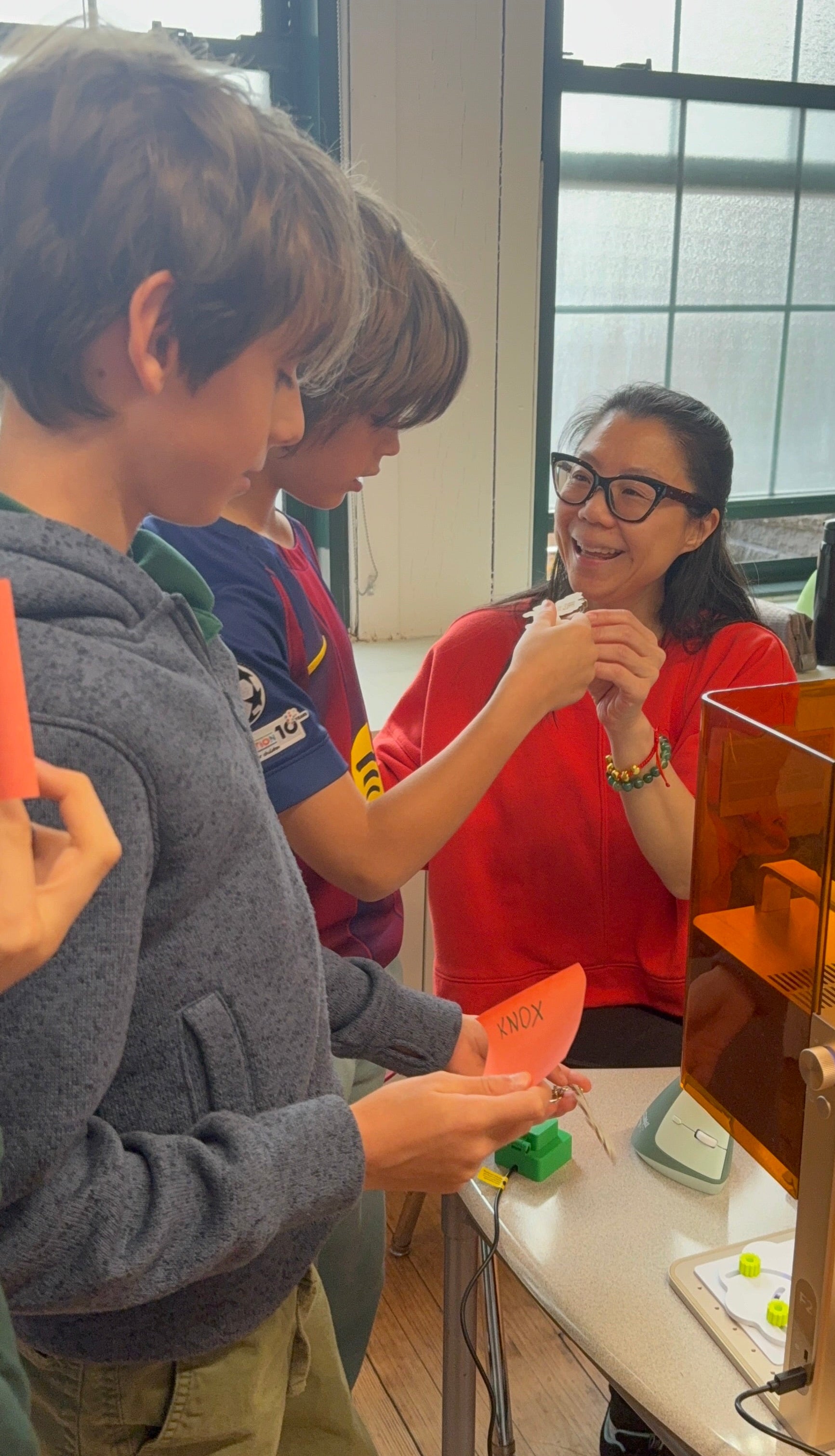 Two children interacting with a woman in front of a laser engraver in a classroom setting.