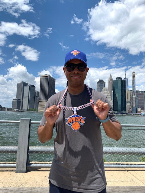 Person wearing a gray shirt with a Knicks hat,  with a city skyline in the background wearing an adult size New York Knicks oversized fan accessory hype chain around his neck.
