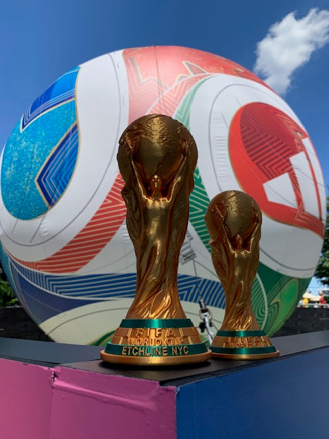 Large and medium gold World Cup trophy 3D prints displayed in front of a soccer ball backdrop
