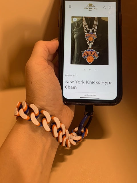 Hand wearing a matching New York Knicks colors Hype Chain wristlet connected with a blue o-ring carabiner. The cell phone screen shows the EtchlineNYC.com website page of the New York Knicks Hype Chain.