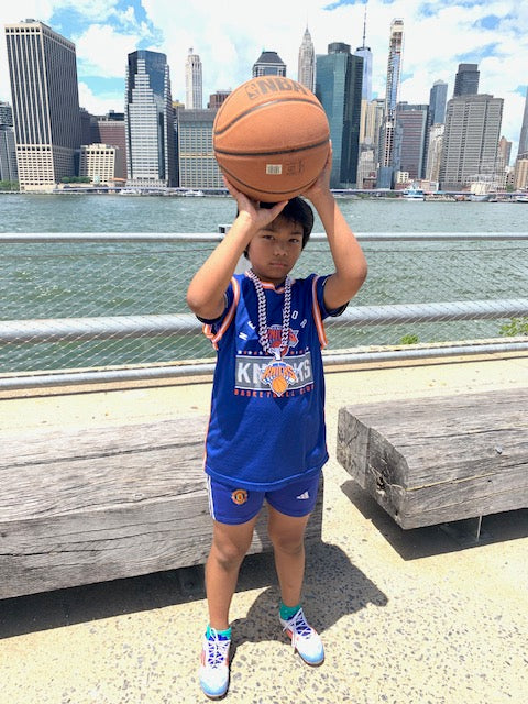 A boy holding a basketball with a city skyline in the background wearing a kids New York Knicks oversized fan accessory hype chain around his neck.