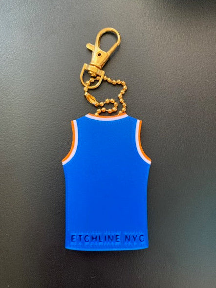Back of personalized Knicks jersey keychain with Etchline NYC maker mark with a gold keychain clasp and chain, resting on a dark gray surface.