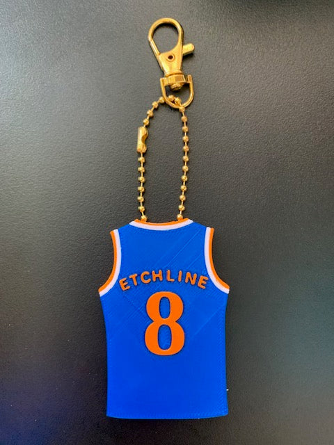 A Knicks soccer jersey-shaped keychain or bag charm with orange lettering reading 'ETCHLINE' and the number 8, attached to a gold-tone ball chain and clasp, shown on a dark gray background.