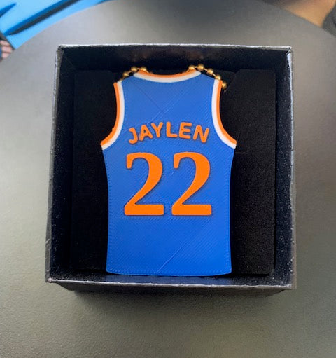 A Knicks basketball jersey-shaped bag charm/ keychain with orange lettering reading 'JAYLEN' and the number '22', displayed inside a black gift box on a gray surface.